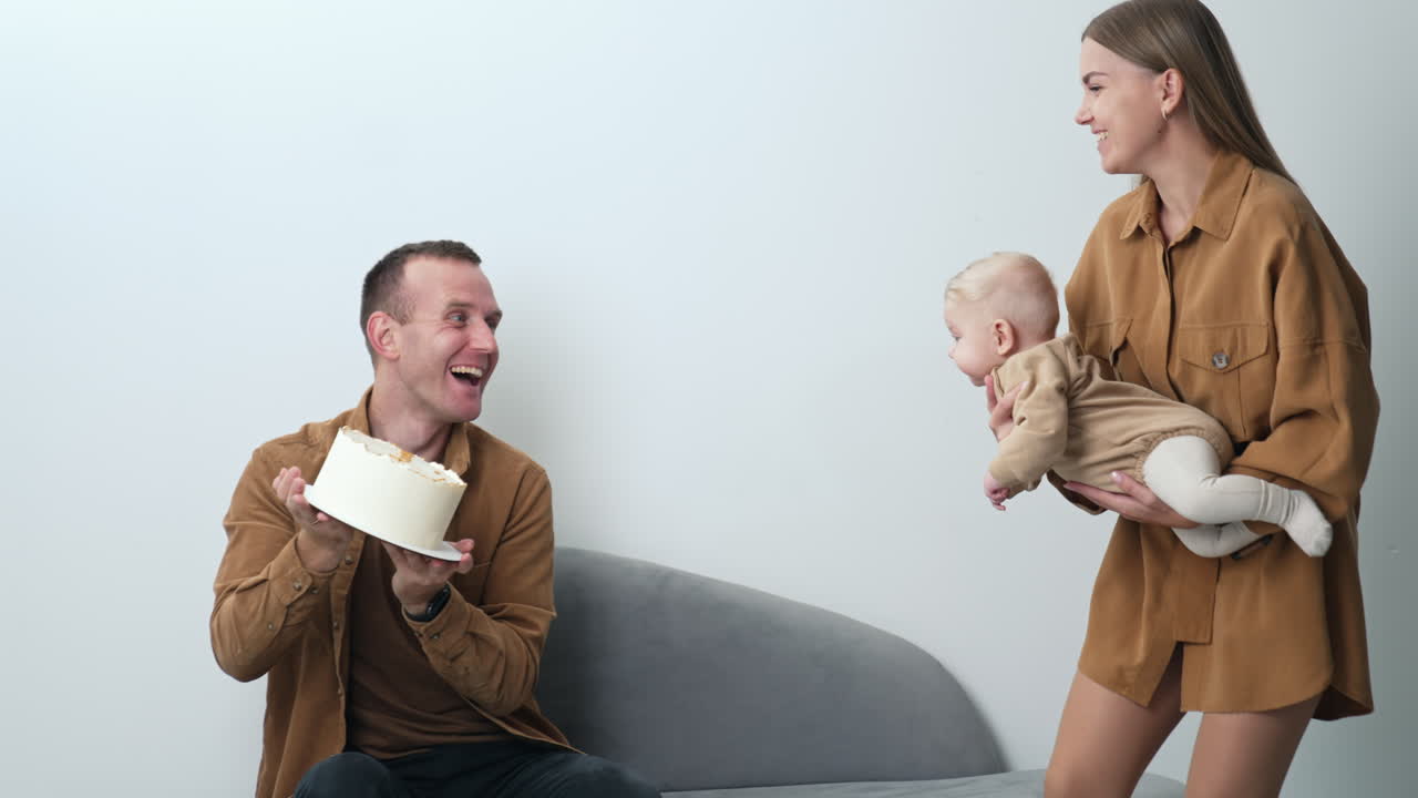 Joyful Caucasian parents amusing their little baby. Mother holds her child taking him closer to a cake in dad's hands.