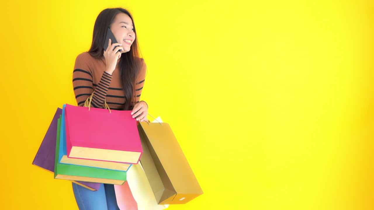 Portrait of happy Asian female with colorful shopping bags and smartphone, Isolated on yellow background