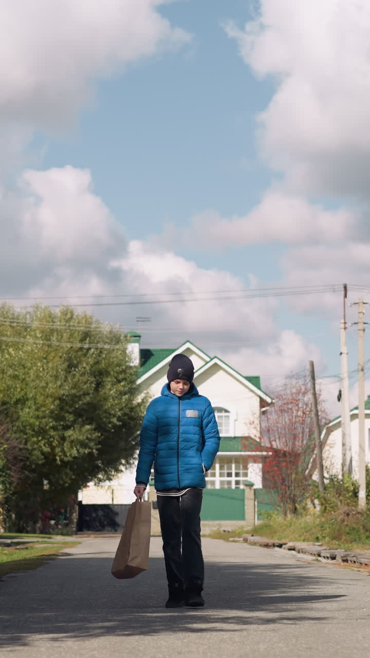 adolescente con capucha azul y gorra negra lleva una bolsa de papel, paseando por la calle suburbana alineada con follaje de otoño y casas distintivas bajo un cielo nublado