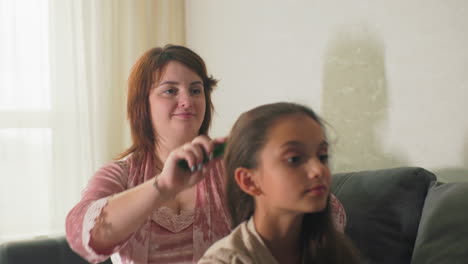 Mother brushing daughter’s hair on couch in cozy living room. Intimate, loving moment between parent and child with soft light from window, creating a calm, peaceful atmosphere in home