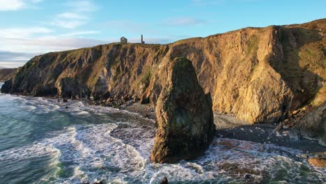 Drone-circling-round-a-massive-sea-stack-at-sunset-on-the-Copper-Coast,-Tankardstown-Copper-Mine-can-be-seen-at-the-cliff-top