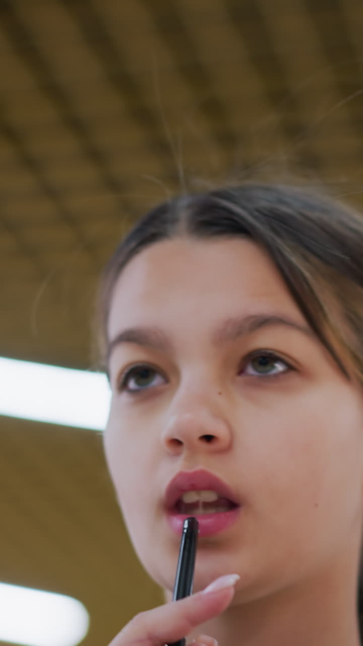 Teen girl with green cup gazing upward indoors, Young girl in plaid jacket drinks from green cup indoors, Teen girl in casual plaid jacket looks upward at ceiling lights with quiet curiosity