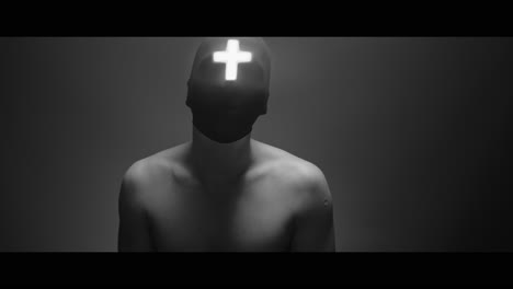 masked and hooded man with cross of light glowing on his face looks in the camera