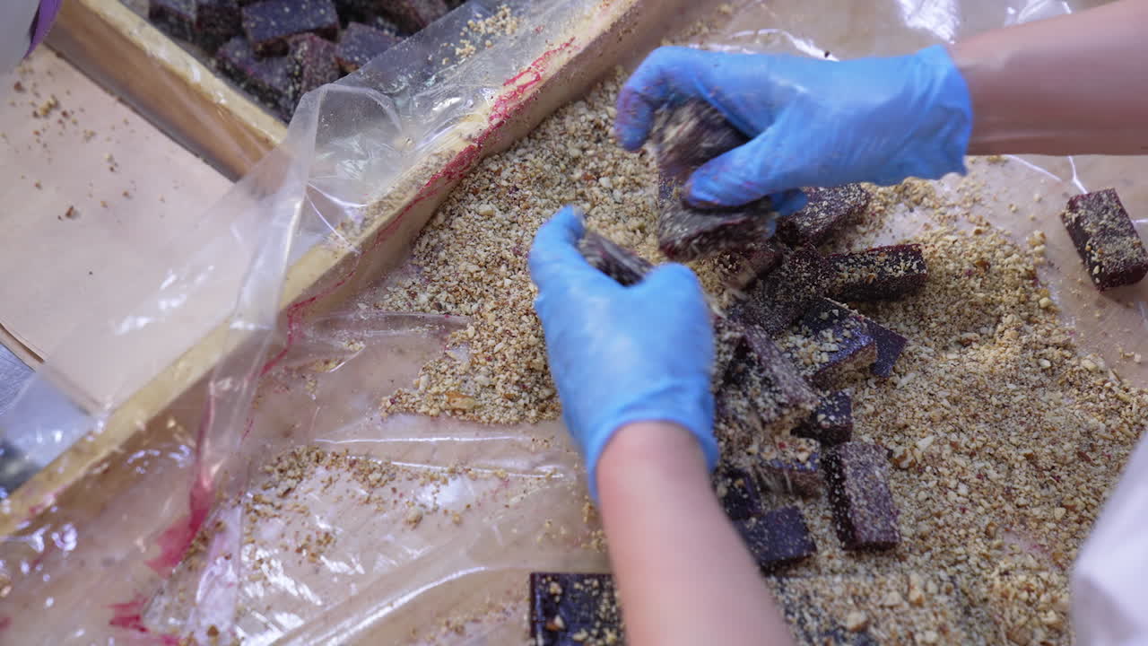 Dark marmalade pieces being coated with crushed nuts. Worker's hands mix up sweet desserts with ground walnuts. Close up.