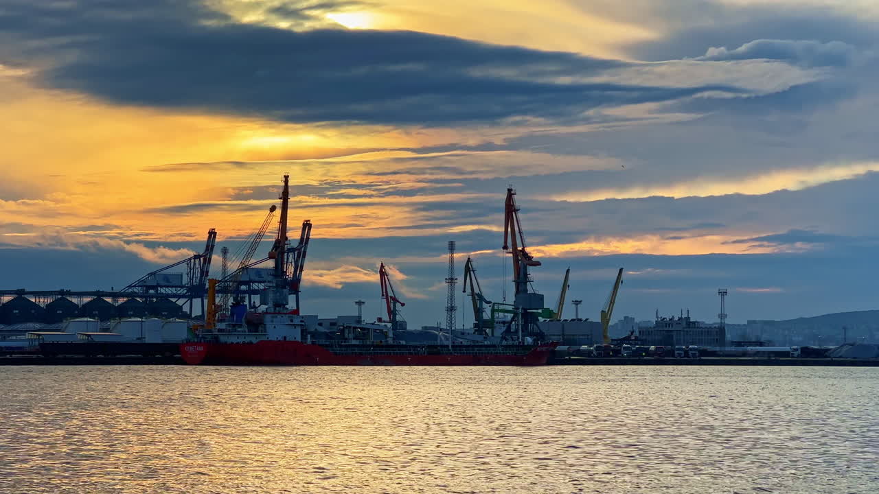 Lastfartyg dockade i hamn under solnedgången med molnig himmel och industrikranar i bakgrunden
