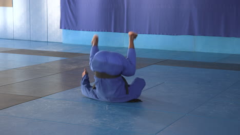 practicing judo, athlete in blue gi rolling on mat in dojo
