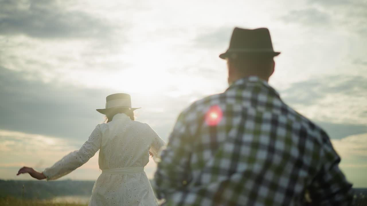 una toma de primer plano de una mujer con un vestido blanco y sombrero siendo perseguida por un hombre con una camisa a cuadros durante la puesta de sol bajo un cielo nublado. la escena captura un momento dinámico y romántico con el sol