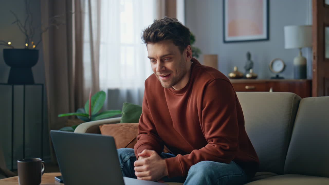 Uomo sorridente in videochiamata con il laptop sul divano dell'appartamento. Il ragazzo ha una riunione online.