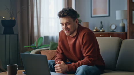 smiling man video calling by laptop at apartment couch. guy have online meeting