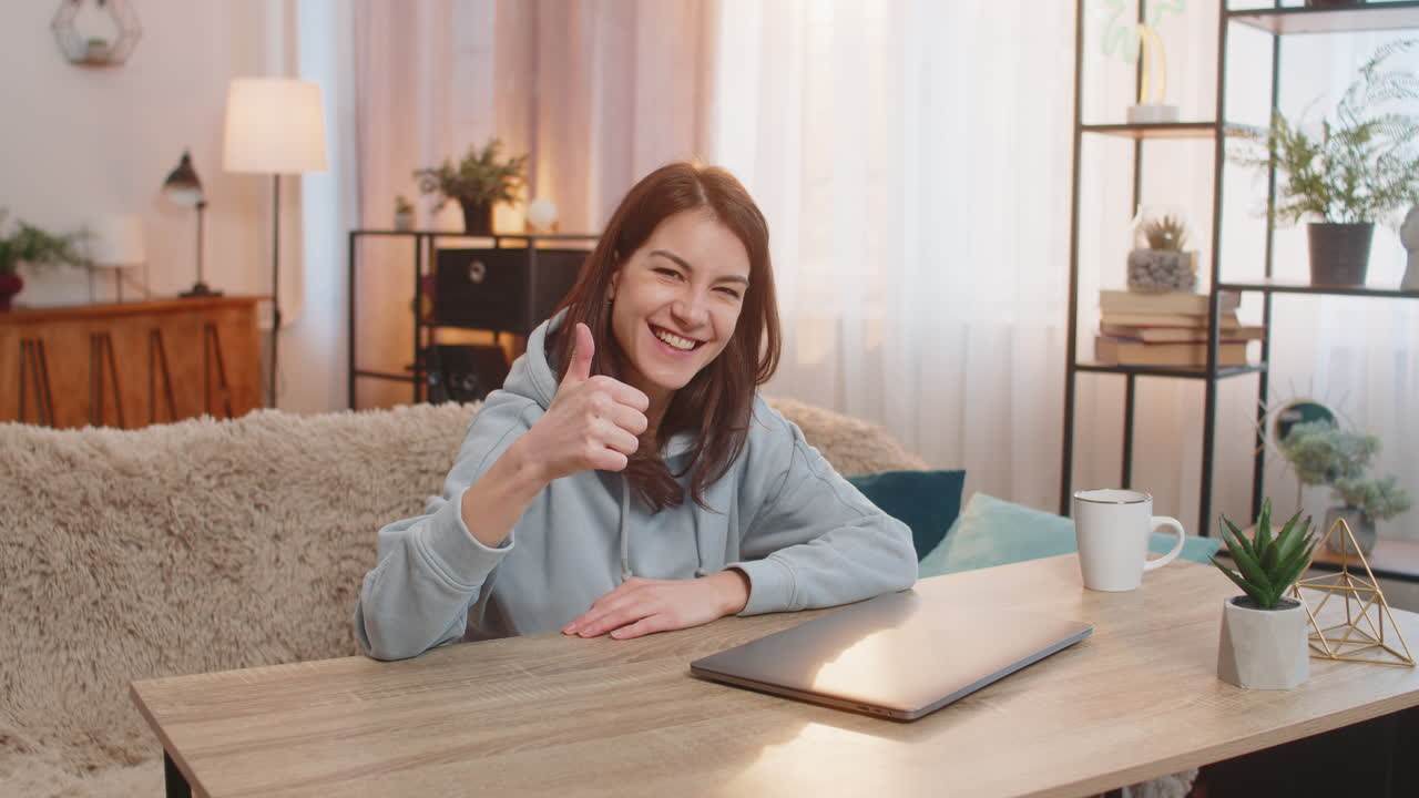 Smiling young woman freelancer closing laptop after work and showing thumbs up at table at home