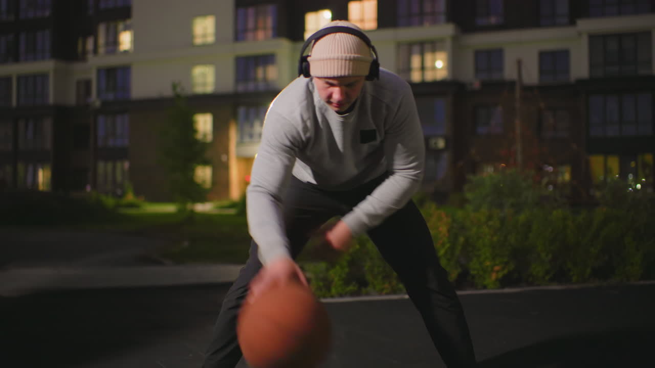 Hvid mand drible basketball om natten i urban gård med hættetrøje hue hovedtelefoner, pickup spiller der øver crossovers, sen aften træner der finjusterer boldkontrol, nærbilleder af hænder