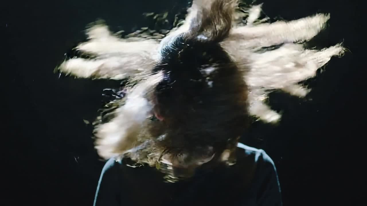 Donna in studio con un top scuro, che scuote rapidamente la testa da una posa piegata, creando archi di capelli lunghi illuminati dal bordo.