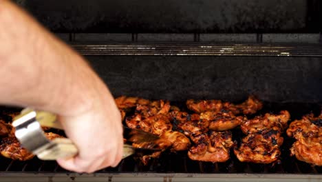 control de brazo-mano de los hombres y volteo de pollo a la barbacoa con pinzas de metal y madera en una parrilla de gas mientras se encienden las llamas - todavía disparo de trípode