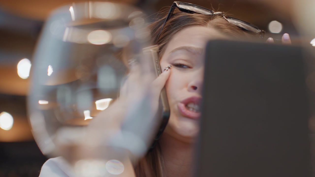 White woman in cafe on phone, young white woman behind laptop seen through glass and bokeh lights, rubbing her eye and holding phone to ear during tense client call sunglasses on head, manicured