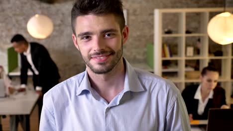 young businessman is watching at camera in office, smiling, his colleagues are networking with technologies, working concept, business concept, communication concept, blurred background