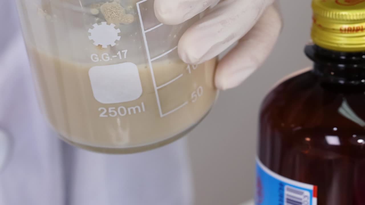 A gloved hand stirs a brown liquid in a 250ml beaker next to a brown bottle.