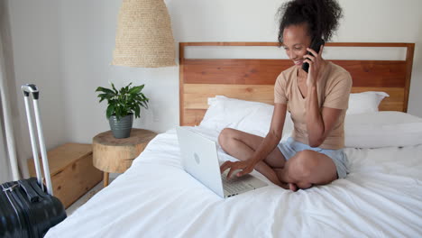 young woman on bed using laptop and talking on phone, suitcase nearby, at home