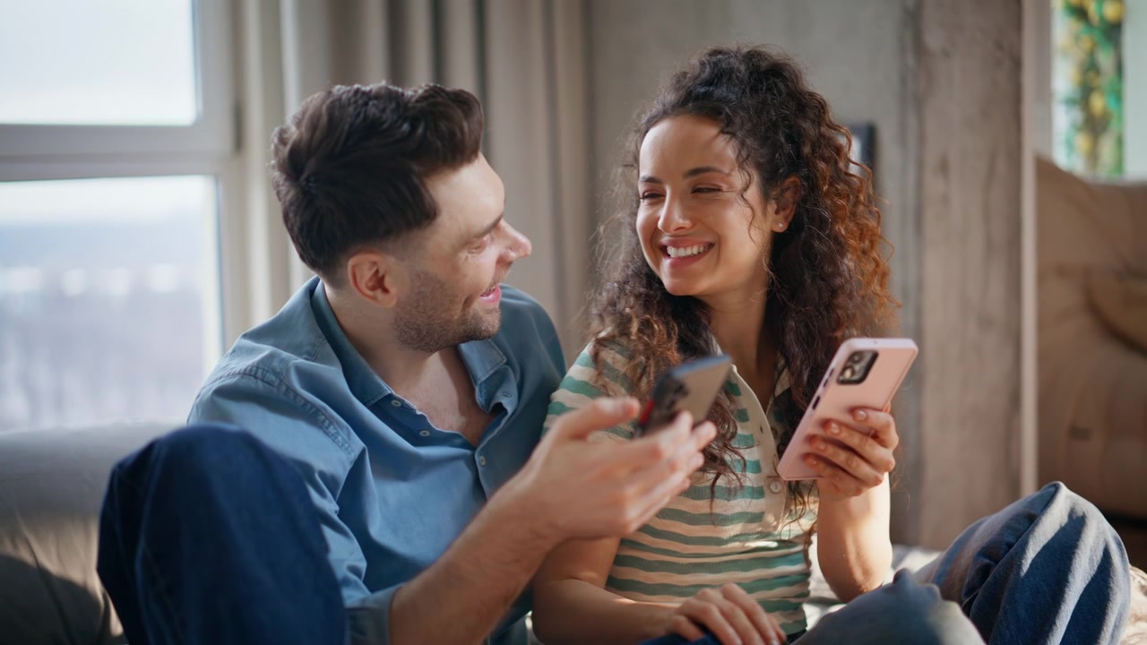 Casal usando celulares juntos, rindo no sofá de casa. Lazer aconchegante de fim de semana