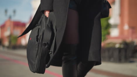 close-up of stylish woman walking confidently on sunlit urban street, wearing black coat and leather boots, carrying two bags, with leg revealed under coat, background features blurred building