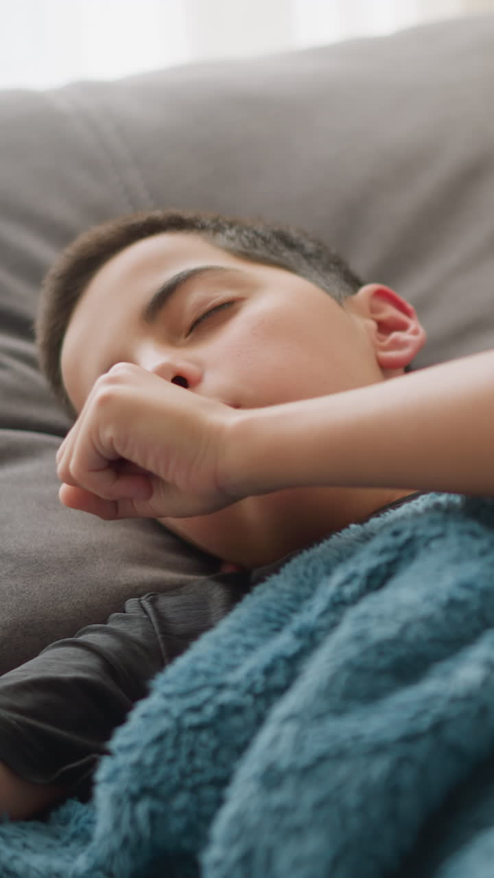 Close-up of child covered with duvet coughing, with tissue near head and light streaming through the window, the child is resting, feeling unwell