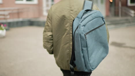close up rear view boy wearing hoodie jacket walking toward school building with large backpack, blurred surroundings of campus entrance creating focus on student movement and academic atmosphere