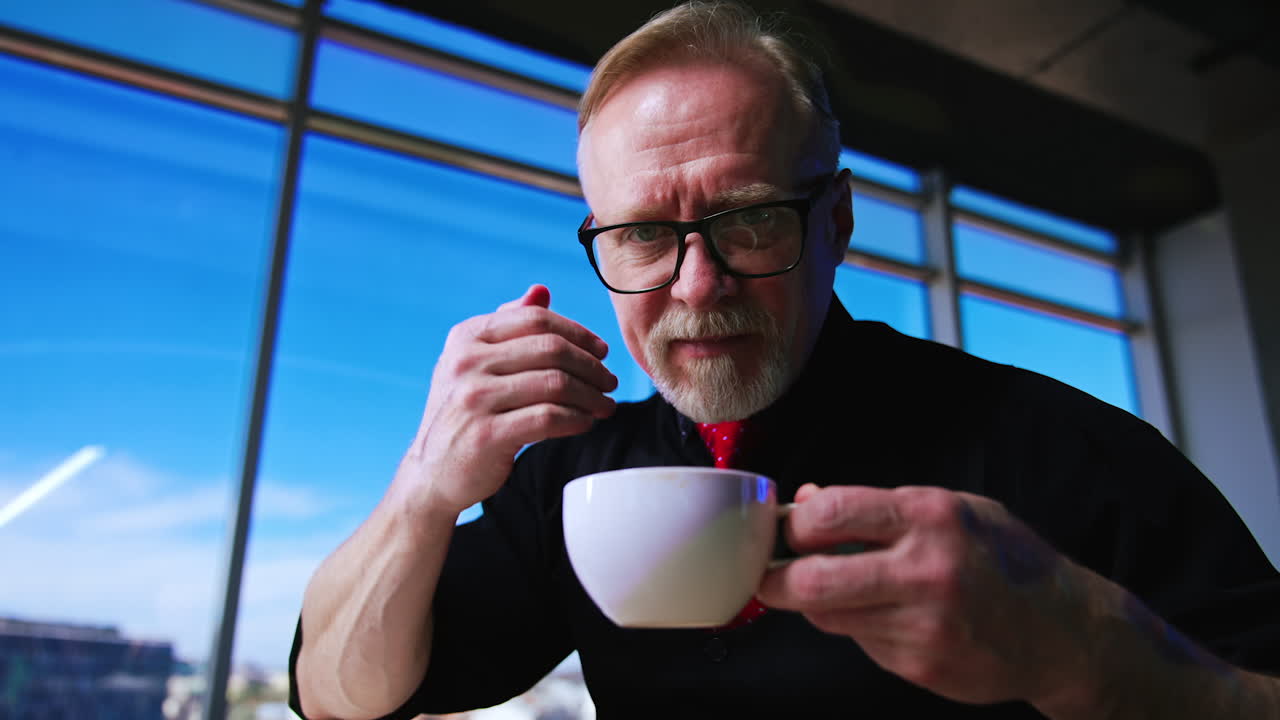 Adult bearded man in glasses waves his hand over the cup trying to spread the coffee flavor. Low angle view on a man holding a cup. Close up.