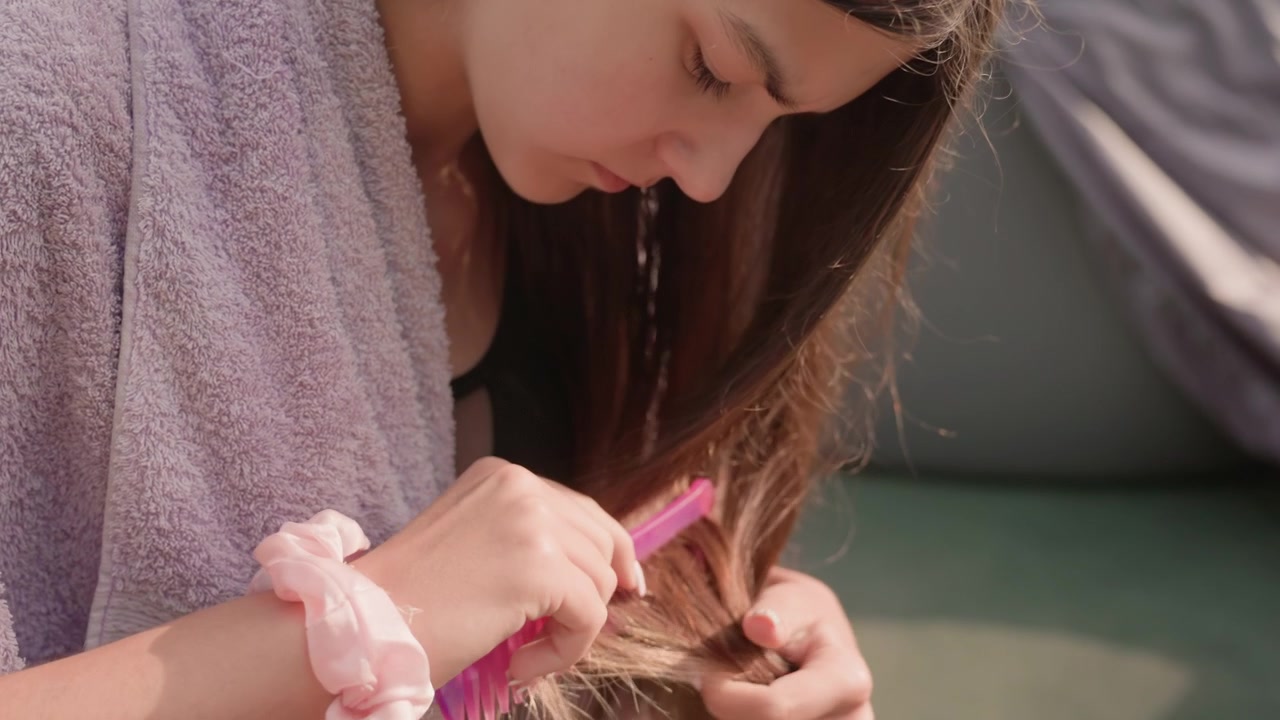 Young girl combs her wet hair with pink comb while wearing purple towel and light pink scrunchie focusing intently on grooming process in outdoor setting