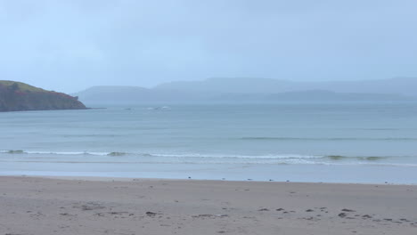 gentle waves on a sandy beach with misty hills in the distance, serene atmosphere, cloudy irish coastline