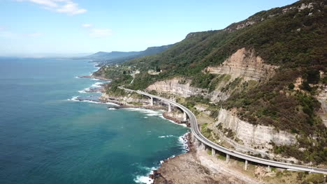 aerial: cars driving along a curving highway next to a stunning ocean coastline, in new south wales australia