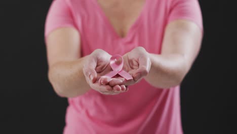 Mid-section-of-woman-holding-a-pink-ribbon-against-black-background