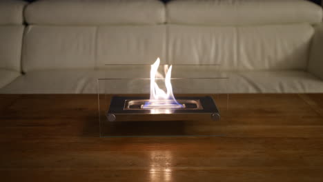 an indoor glass fronted fireplace in a living room on a table with a sofa in the background of the room