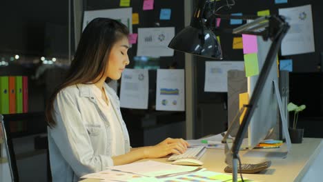 beautiful asian female office worker works late hours. she's alone on the office. her table is lit by a lamp