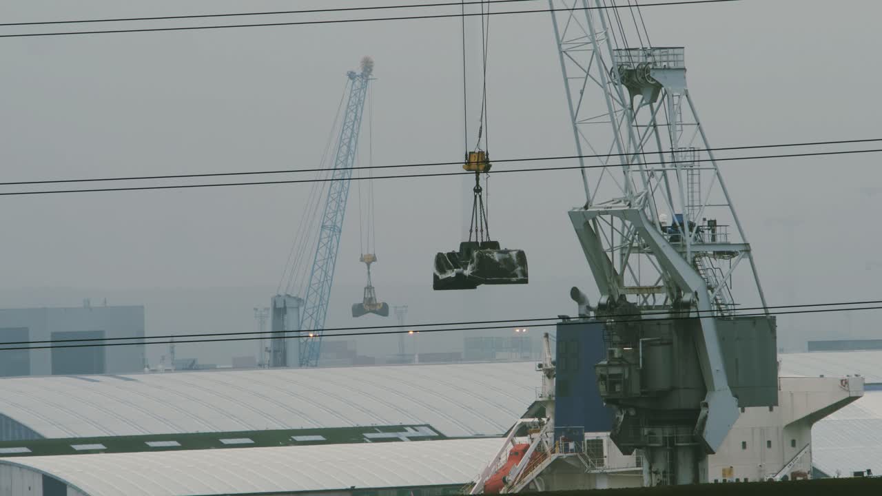 Stationary industrial cranes subtly move in the wind at a misty harbor under an overcast sky, with structural details creating a calm yet atmospheric maritime and industrial scene