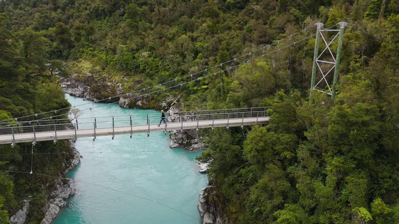 Breathtaking static overview of Hokitika Gorge with turquoise water flowing through lush, forested cliffs in New Zealand as solo traveler walks along bridge