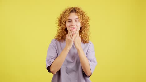 a woman with curly blonde hair blowing kisses and smiling against a yellow background