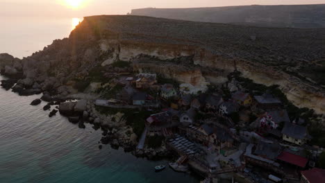 Vista-Aérea-Del-Colorido-Sol-Vespertino-Detrás-Del-Pueblo-De-Popeye,-En-Malta