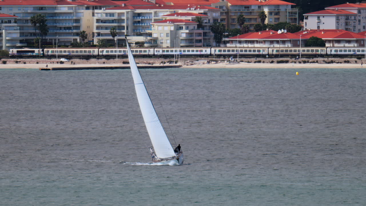 Juan-les-Pins, France - April 3, 2025: Sail boat moving on the sea with a train moving on the rails in the city on the background