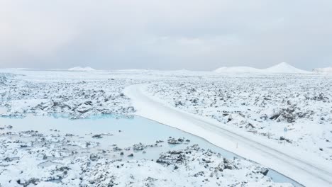 Winding-road-covered-in-snow-with-blue-geothermal-pool-next-to-it,-Iceland