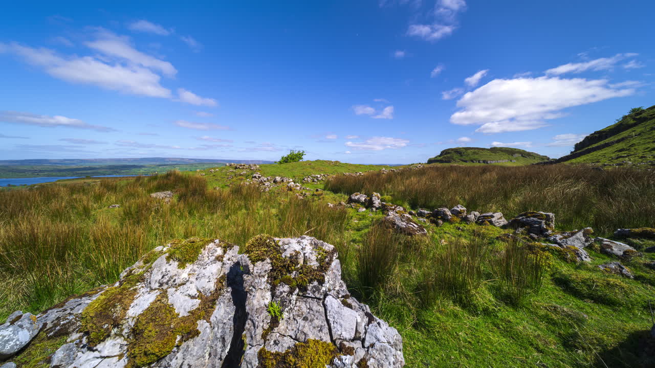 Panorama-timelapsevideo af et landligt landskab med klippeformation i græsset i forgrunden og bakker i det fjerne punktet set fra Carrowkeel i County Sligo i Irland på en solrig dag med skyer.