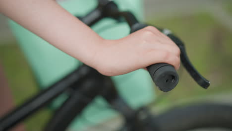 primer plano de una persona agarrando el manillar de una bicicleta, acompañada de una bolsa de color menta, mientras camina por un camino en un parque sereno con vegetación borrosa en el fondo