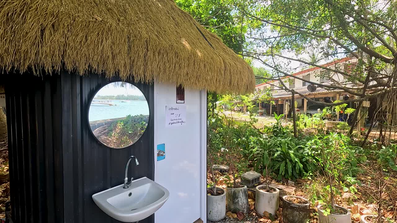 A rustic outdoor toilet with a thatched roof amidst lush greenery in Phuket, Thailand, captured in bright daylight