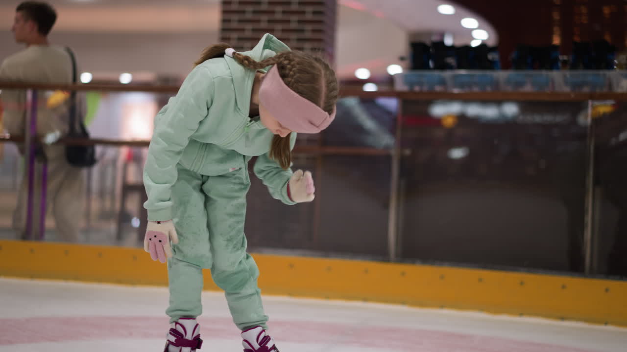 una vista cercana de un niño en un traje verde menta y una cinta rosa inclinándose para limpiarse en una pista de hielo, el fondo muestra una vista borrosa de otras personas y equipo de patinaje sobre hielo