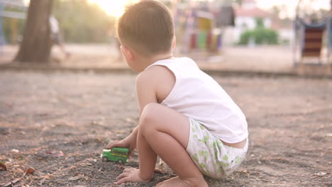 Zeitlupenvideo-Eines-Süßen-Kleinen-Asiatischen-Jungen,-Der-Allein-Mit-Dem-Dreck-Und-Autos-Und-älteren-Kindern-Spielt,-Die-Im-Hintergrund-Zusammen-Spielen