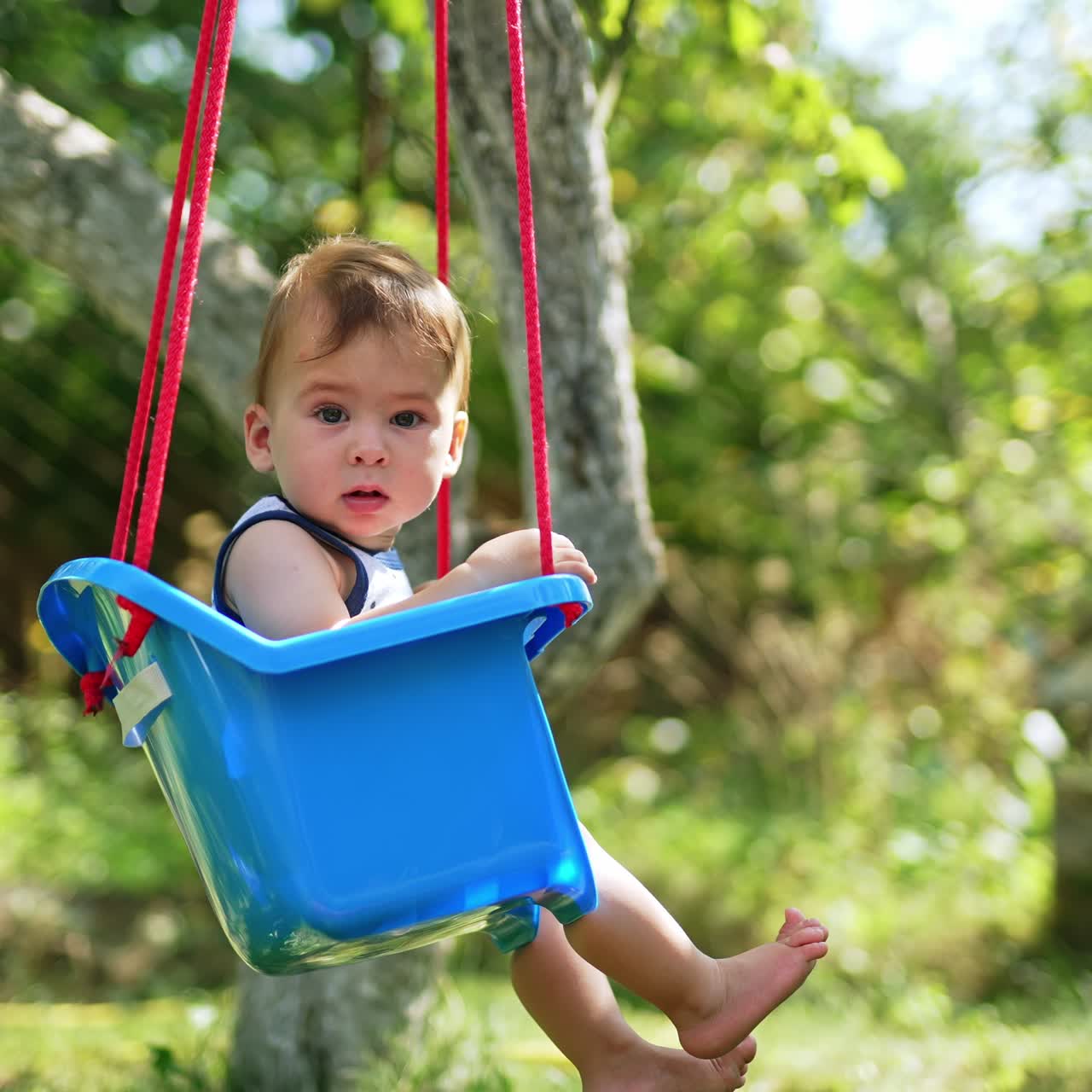 Lovely toddler swinging on a blue swing in the garden. Baby looking attentively into camera. Side view. Blurred backdrop