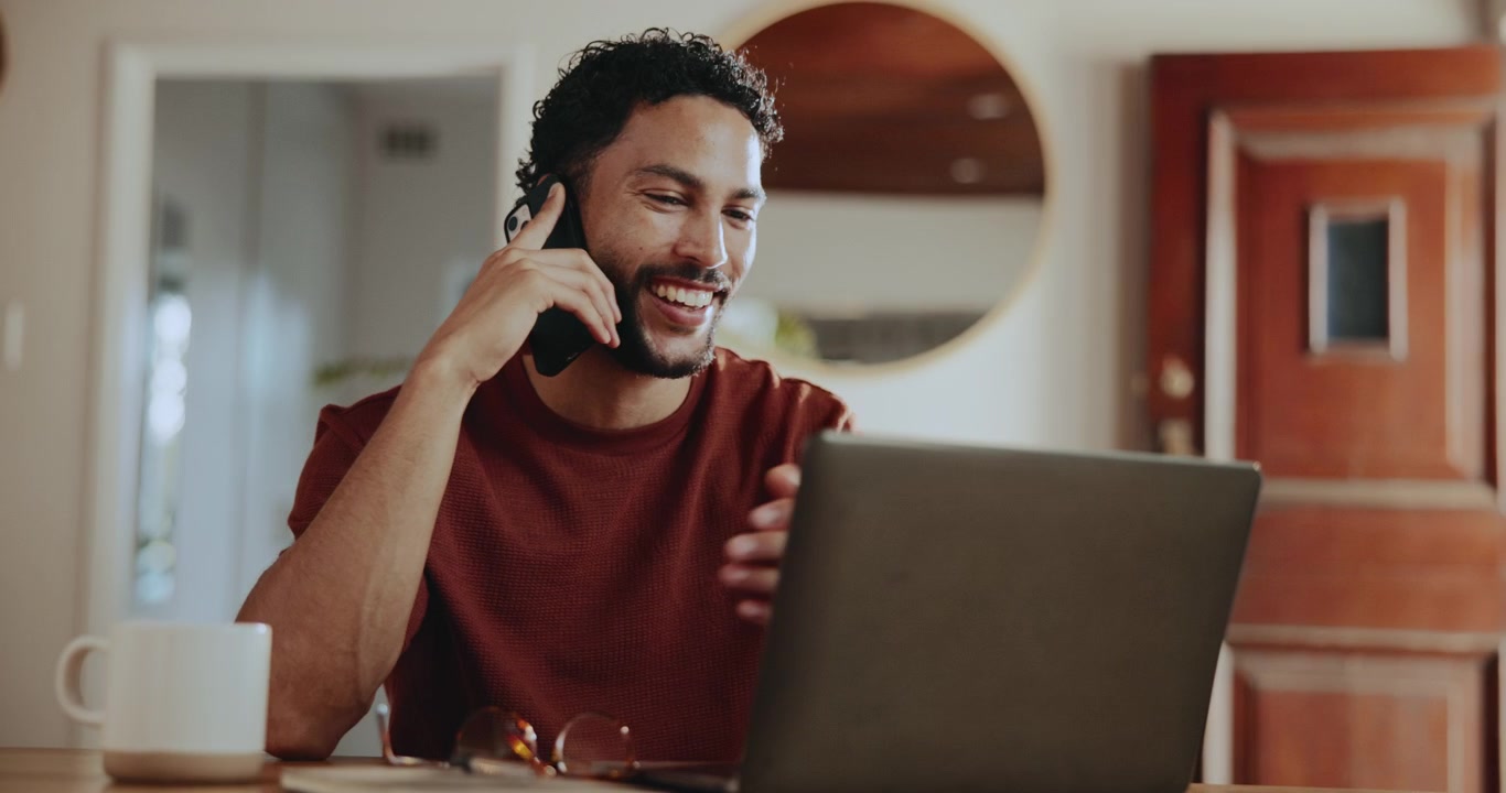 Ung mand arbejder på bærbar computer og taler i telefon derhjemme
