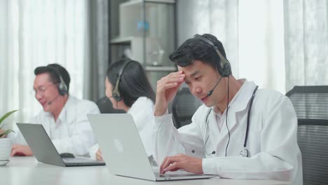 a man of three asian doctors with stethoscopes wearing headsets working as call center agent is tired due to working while his colleagues are talking during working at the office
