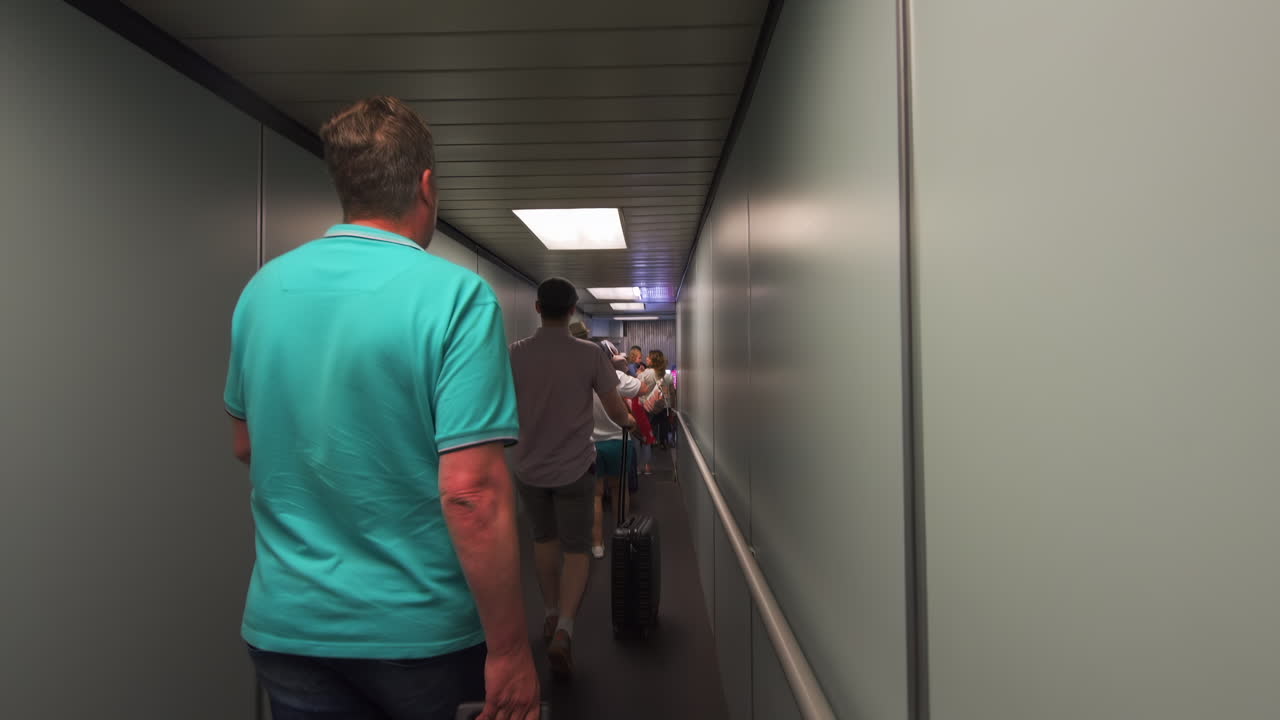 A traveler wearing a blue shirt walks with a suitcase down the jet bridge at an airport, preparing to board a flight. The surrounding area is modern and well-lit