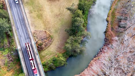 overhead drone footage shows trucks and a car driving across a bridge above a winding river in rural coonabarabran, australia, under soft daylight
