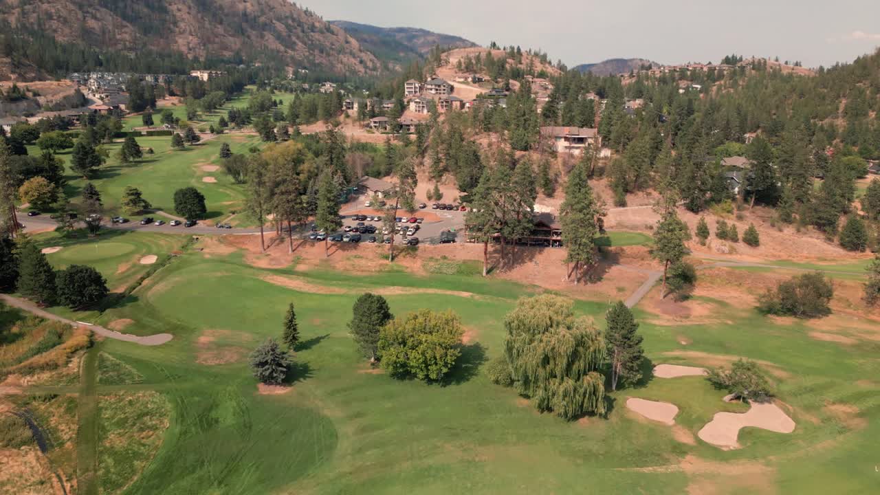 High flying Aerial forward panning down onto Shannon Lakes Golf course West Kelowna Okanagan Valley Green yellow grasses lush trees vibrant greenery foliage mountain range blue sky light clouds day