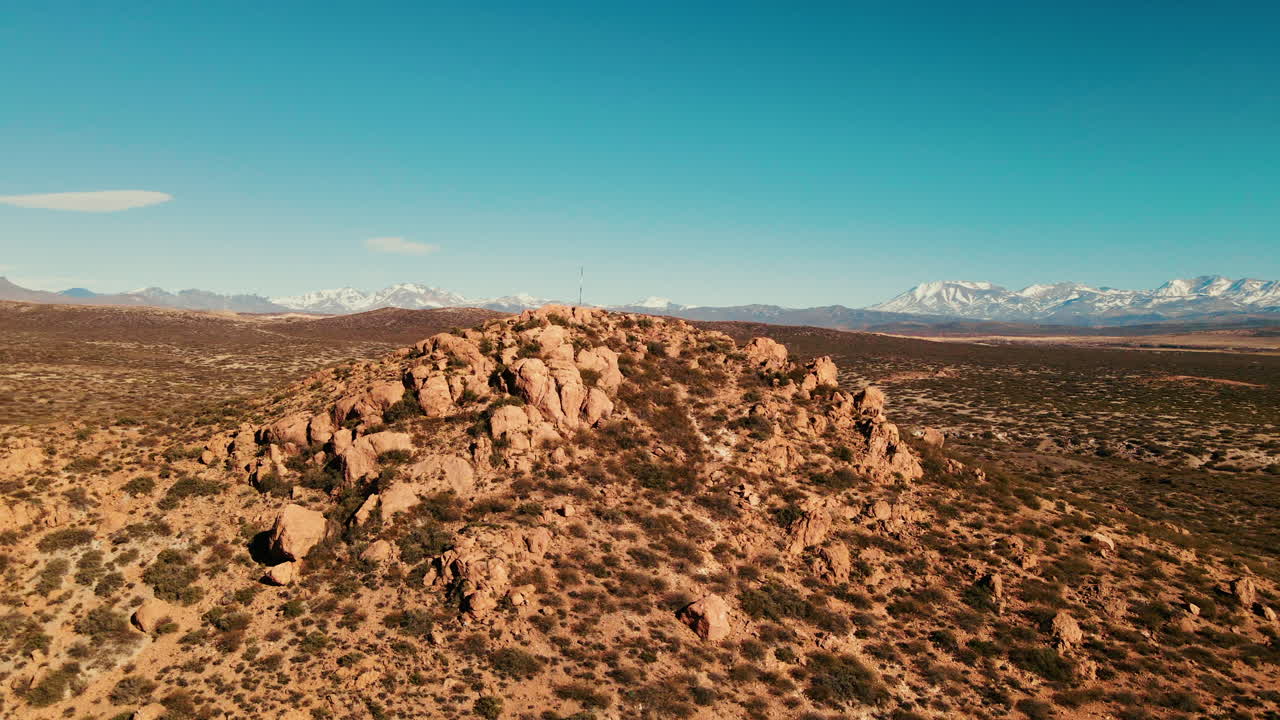 Drone circling around a small mountain crowned with a cross, unveiling the town of Malarg&uuml;e and the Andes mountain range in the background
